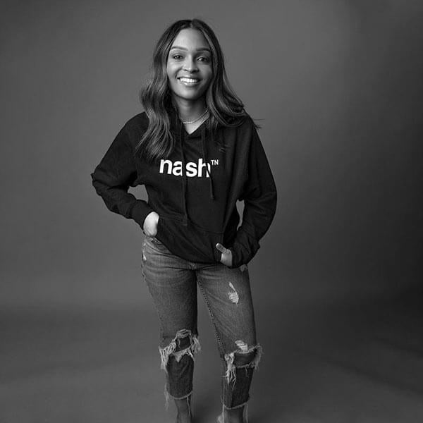 Lady standing in front of a gray background with hands in pockets of black hooded sweatshirt. She is wearing ripped jeans and smiling. 