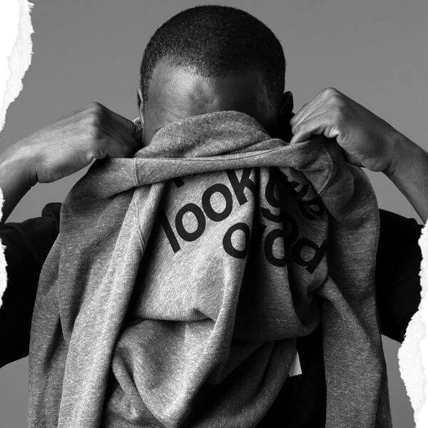 Man with short hair taking off a gray crewneck sweatshirt on a gray background. Black lettering is partially showing on the sweatshirt. The sweatshirt is partially covering his face his hands are holding the shirt neck near his ears.