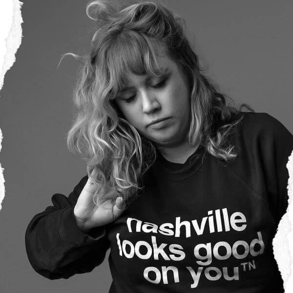 Woman wearing a black sweatshirt with white text on a gray background. She is touching her light colored hair with her right hand. Her sweatshirt has white text and says nashville looks good on youᵀᴺ.  