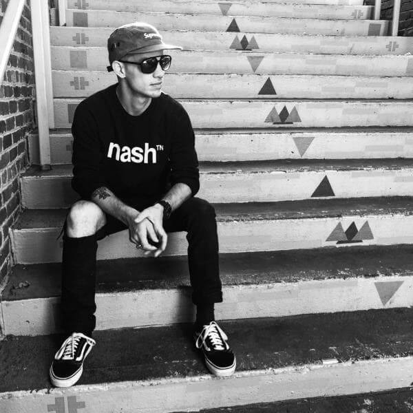 Man sitting on row of stairs with his arms on his knees and hand clasped. He is wearing black pants with a hole in the knee, black nashᵀᴺ  sweatshirt with white logo, a hat and sunglasses.
