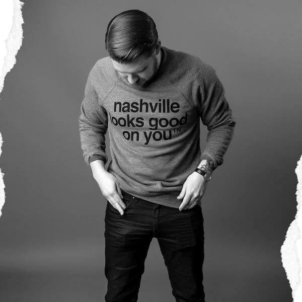 Man wearing black pants and gray sweatshirt with a gray background. His head is looking down and his hair is combed to the side. His sleeves are rolled up and he is wearing a watch on his left wrist and his sweatshirt has nashville looks good on you nashᵀᴺ in black. 