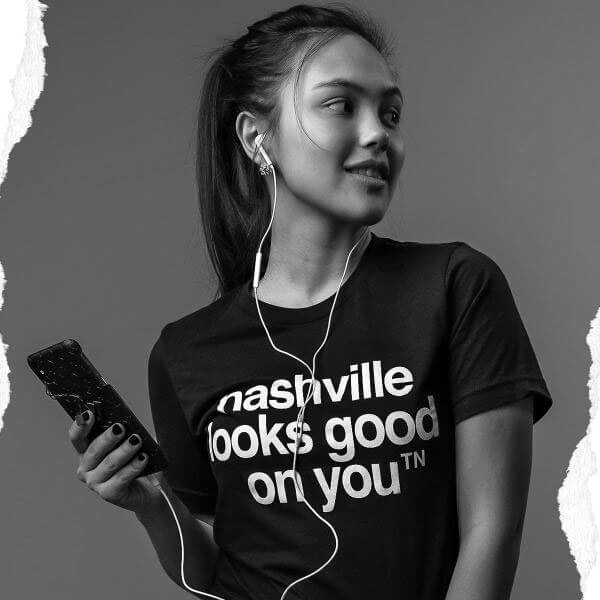 Girl wearing a black T-shirt listening to headphones in front of a gray background. She is wearing a black crewneck short sleeve T-shirt with white text. She is holding her phone with her right hand and looking over her left shoulder. The nashᵀᴺ slogan, nashville looks good on youᵀᴺ is printed in white and centered on shirt.