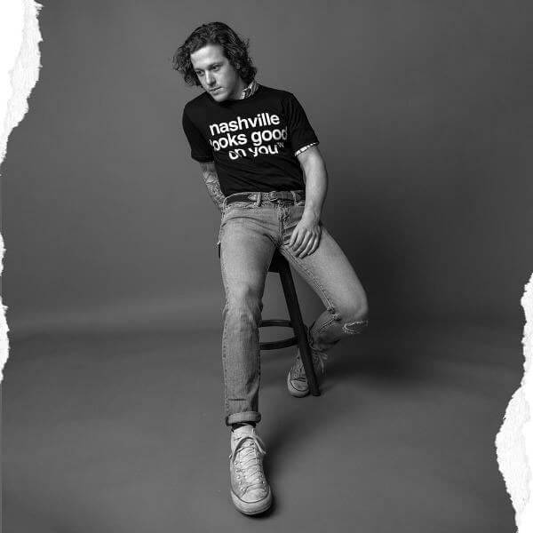 Man sitting on a stool in jeans and black T-shirt in front of a gray background. He is wearing white sneakers and his right leg is closer to the camera and he looks like he is leaning backward. He is wearing a black crewneck short sleeve T-shirt with white text. The nashᵀᴺ slogan, nashville looks good on youᵀᴺ is printed in white and centered on shirt.