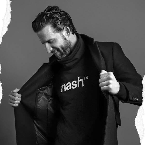 Man with short hair and beard is looking down and opening his jacket to show us his shirt underneath. He is wearing a black, short sleeve T-shirt with white, nashᵀᴺ  logo on the front.