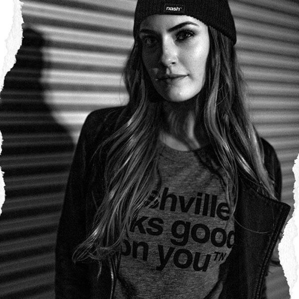 Woman wearing a black jacket, gray shirt and black beanie hat in front of a metal textured roll up door. Her long hair hangs over her shoulders to partially reveal the black shirt text nashville looks good on youᵀᴺ.