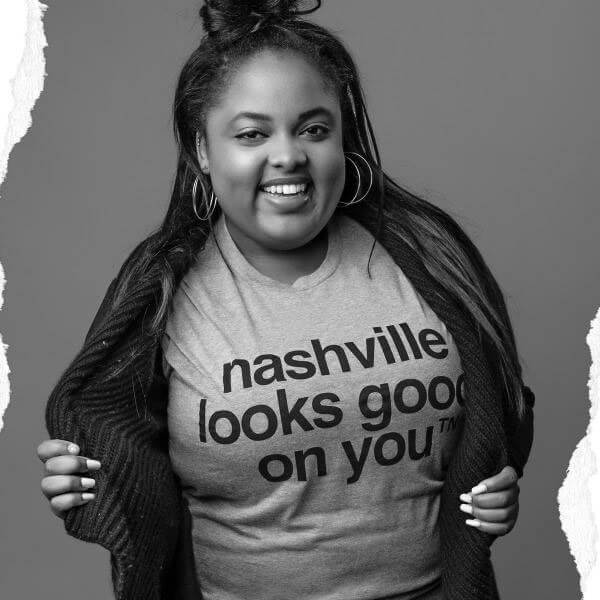 Lady smiling in front of a gray background. Her hair is in a top knot and she has on large double hoop earrings. She has on a dark color cardigan and gray T-shirt with black text. She is holding the edges of her cardigan open to show us what the shirt says. She has long white painted fingernails. Her T-shirt says nashville looks good on youᵀᴺ  in black. 