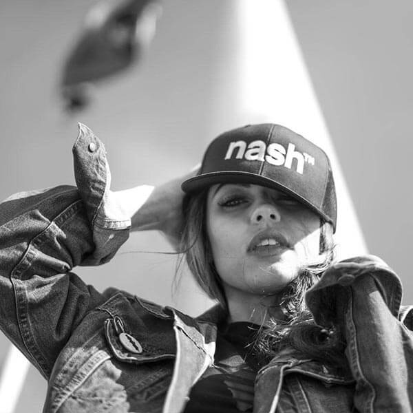 Woman in blue jean jacket and black baseball cap looking down at the camera. Her right hand is on the back of her head and the baseball cap is slightly tilted to the side. The logo nashᵀᴺ  is stitched in white on the front center of the cap.