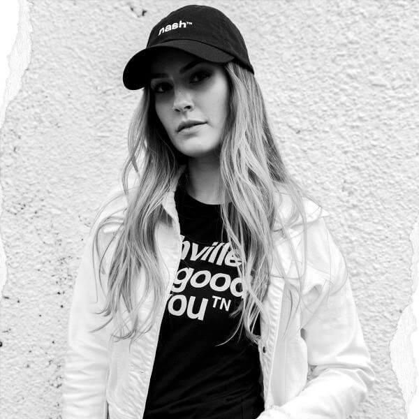 Lady leaning on a white, textured wall. She has long hair draped over the front of her shoulders. She has on a white jacket, black hat with white letters and a black shirt with white letters that says nashville looks good on youᵀᴺ.