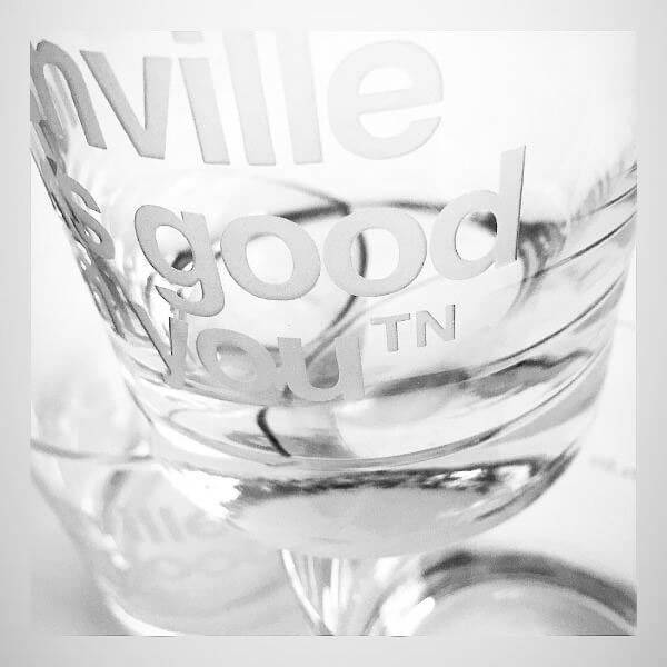 A closeup detail of the etched letters on a clear rocks glass. Clear glass with frosted letters on a white background. On the front center of the glass, nashᵀᴺ slogan nashville looks good on youᵀᴺ is etched.