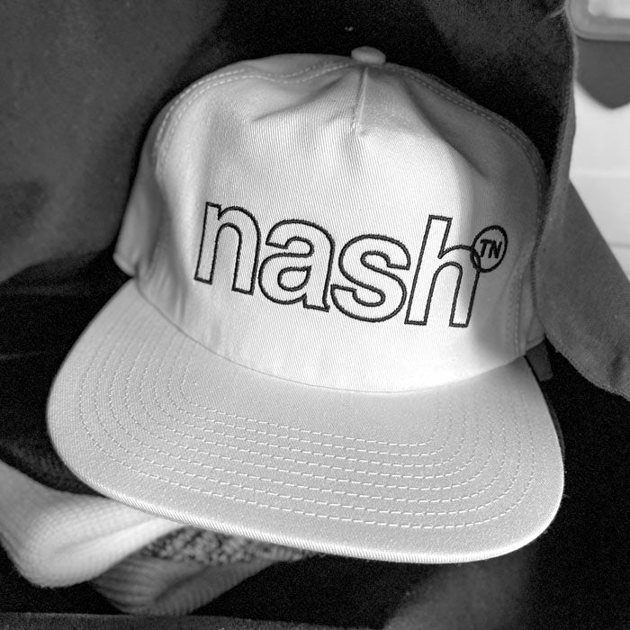 A stack of merchandise shown inside a shopping bag. A white baseball  cap with black outlined letters is shown on top. Nashᵀᴺ is outlined in black and the superscript TN is inside a black, outlined circle.