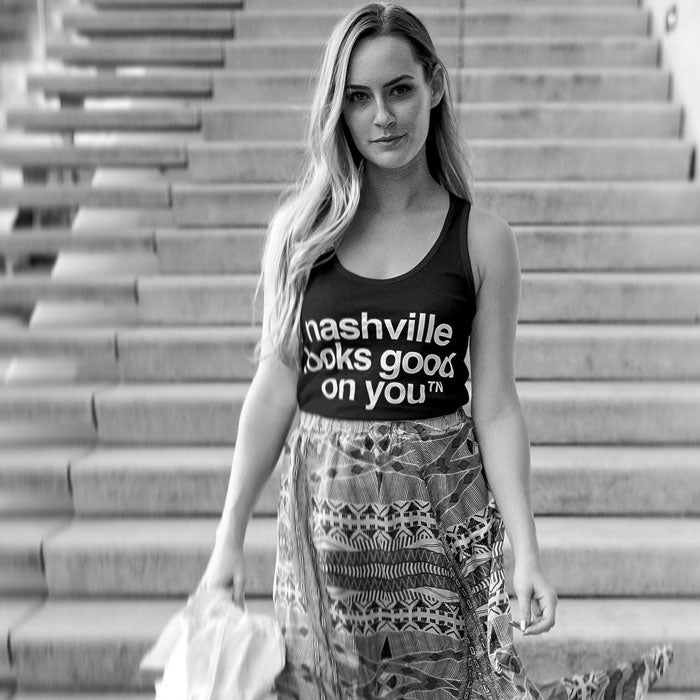 Lady shown in front of tall staircase wearing a black tank top with white letters and patterned long skirt.