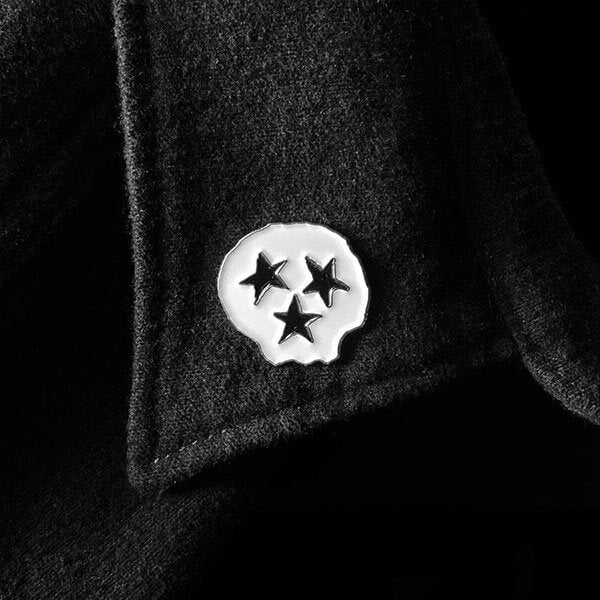 A white, skull shaped fashion pin on a black shirt lapel. The pin has three black stars that represent both the face and Tennessee's Tri-star moniker for the three grand divisions of Tennessee: East, Middle and West.