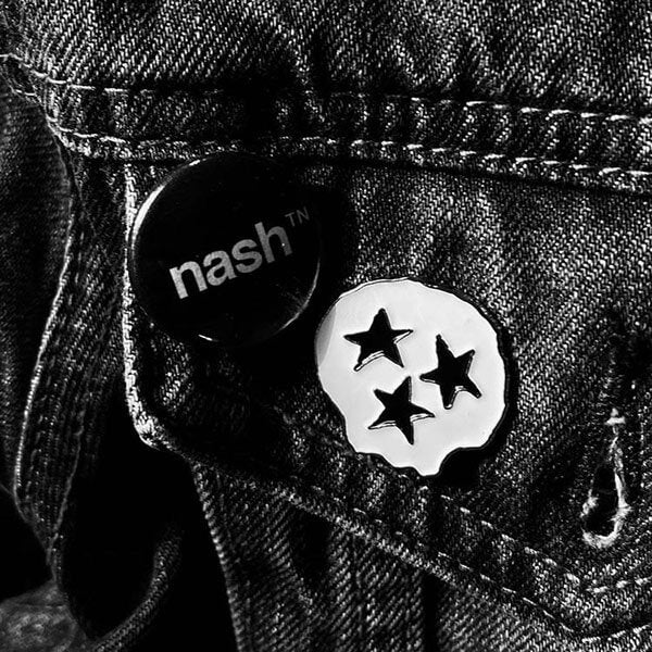 Closeup of two fashion pins on the pocket of a denim jacket. One pin is a white skull shaped pin with star eyes and mouth. The other pin is a black circle with white letters that spell nashᵀᴺ  for Nashville, Tennessee.